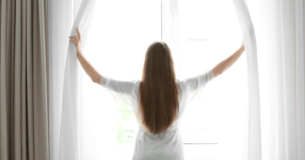 前に向かいカーテンを開ける女性の後ろ姿